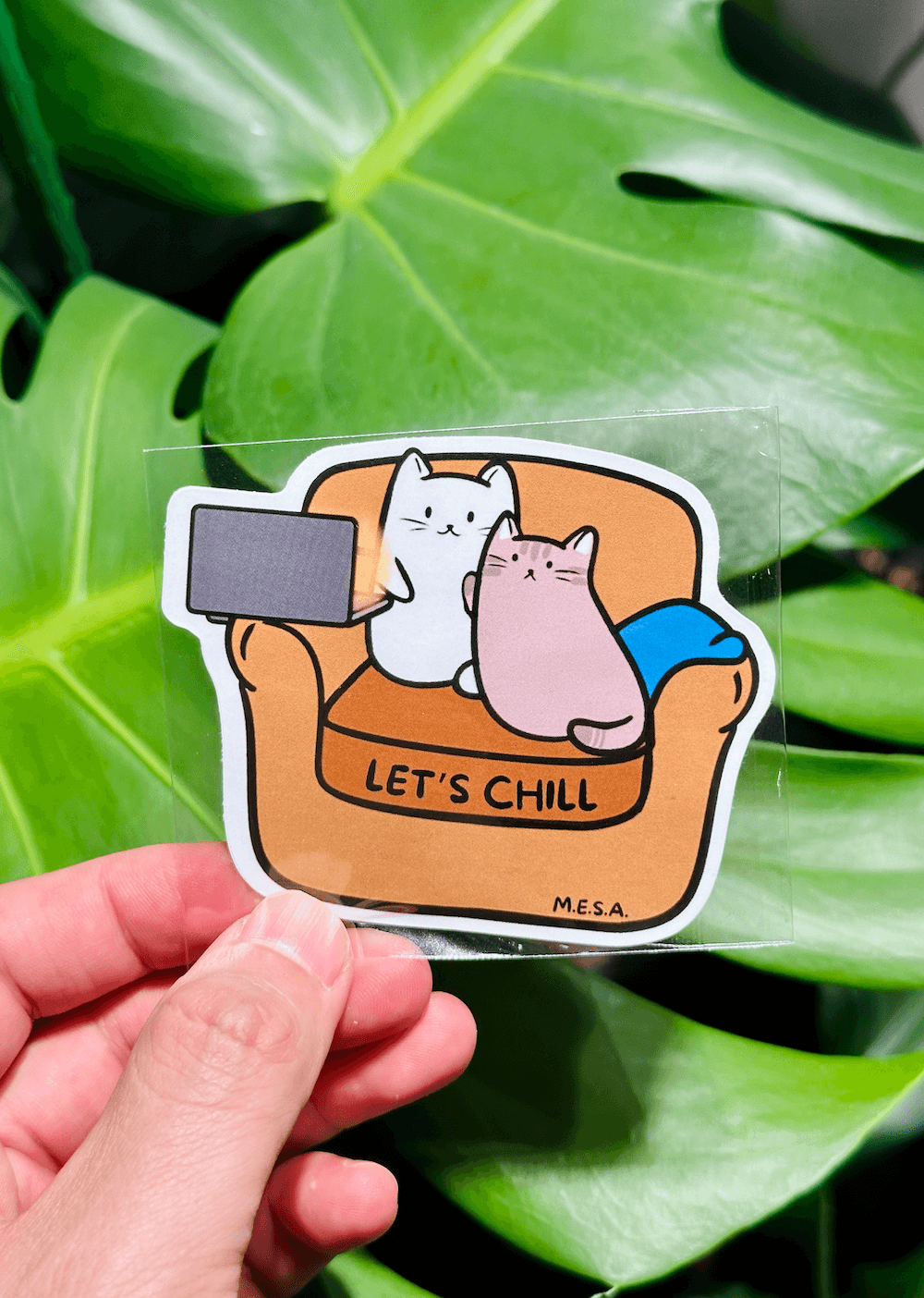 Cute cat sticker with two cats relaxing on a brown couch, text "Let's Chill," set against a green leafy background.